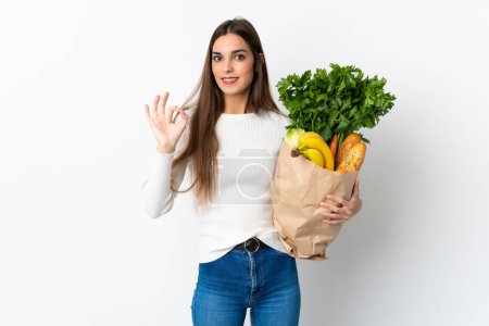 Foto de Joven mujer caucásica comprando algunos alimentos aislados sobre fondo blanco mostrando signo de ok con los dedos - Imagen libre de derechos