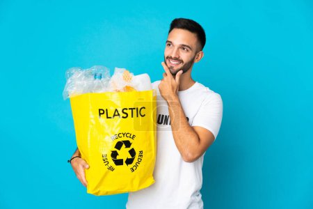 Foto de Joven hombre caucásico sosteniendo una bolsa llena de botellas de plástico para reciclar aislado sobre fondo azul pensando en una idea mientras mira hacia arriba - Imagen libre de derechos