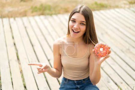 Foto de Adolescente chica sosteniendo un donut al aire libre sorprendido y señalando el dedo a un lado - Imagen libre de derechos