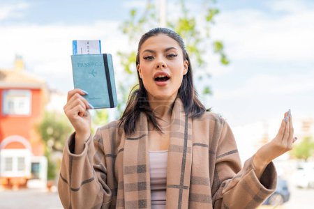 Téléchargez les photos : Jeune jolie femme tenant un passeport à l'extérieur avec une expression faciale choquée - en image libre de droit