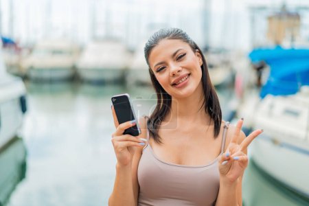 Téléchargez les photos : Jeune jolie femme utilisant téléphone mobile à l'extérieur souriant et montrant signe de victoire - en image libre de droit