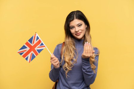 Téléchargez les photos : Jeune fille russe tenant un drapeau du Royaume-Uni isolé sur fond jaune invitant à venir avec la main. Heureux que tu sois venu - en image libre de droit