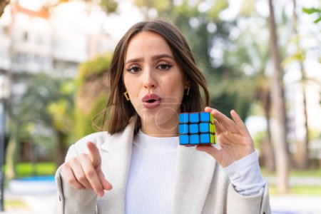 Téléchargez les photos : Jeune jolie femme tenant un cube de puzzle tridimensionnel à l'extérieur surpris et pointant devant - en image libre de droit