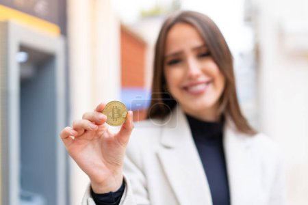 Téléchargez les photos : Jeune jolie femme tenant un Bitcoin à l'extérieur avec expression heureuse - en image libre de droit