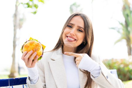 Foto de Joven mujer bonita sosteniendo una hamburguesa al aire libre y señalándolo - Imagen libre de derechos