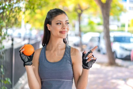 Photo for Young pretty sport woman holding an orange at outdoors pointing to the side to present a product - Royalty Free Image