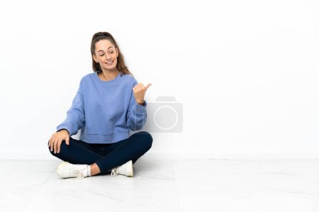 Foto de Mujer joven sentada en el suelo apuntando hacia un lado para presentar un producto - Imagen libre de derechos