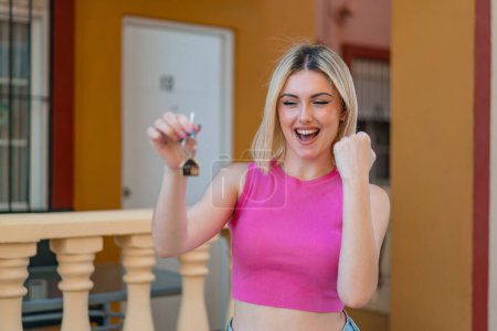 Téléchargez les photos : Jeune jolie femme blonde tenant les clés de la maison à l'extérieur célébrant une victoire - en image libre de droit