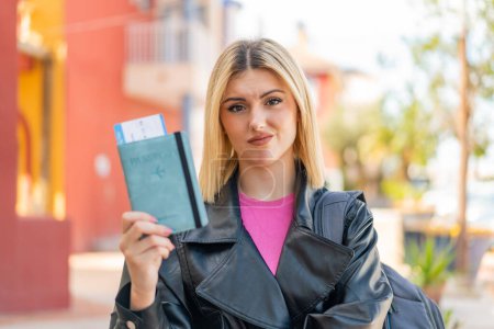 Téléchargez les photos : Jeune jolie femme blonde tenant un passeport à l'extérieur avec une expression triste - en image libre de droit