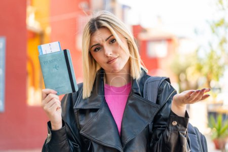 Téléchargez les photos : Jeune jolie femme blonde à l'extérieur tenant un passeport et ayant des doutes - en image libre de droit