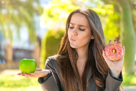 Foto de Mujer joven al aire libre sosteniendo manzana y donut y pensando - Imagen libre de derechos