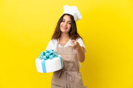 Téléchargez les photos : Jeune chef pâtissier caucasien femme avec un gros gâteau isolé sur fond jaune pointant devant avec expression heureuse - en image libre de droit