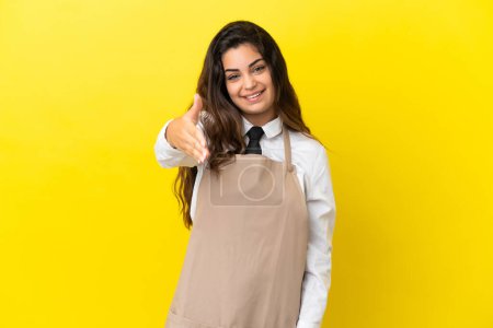 Téléchargez les photos : Jeune serveur restaurant caucasien isolé sur fond jaune serrant la main pour fermer une bonne affaire - en image libre de droit