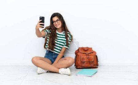 Téléchargez les photos : Jeune étudiante femme assise sur le sol isolé sur fond blanc faisant un selfie - en image libre de droit