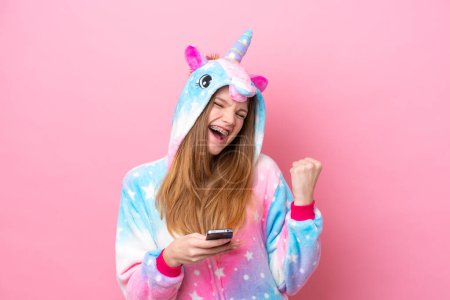 Foto de Adolescente chica rusa con pijama de unicornio aislado sobre fondo rosa con teléfono en posición de victoria - Imagen libre de derechos