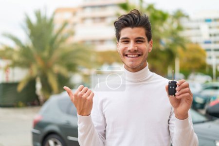 Téléchargez les photos : Jeune homme caucasien tenant des clés de voiture à l'extérieur pointant vers le côté pour présenter un produit - en image libre de droit