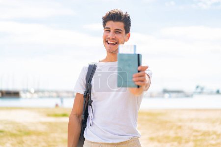 Foto de Joven caucásico sosteniendo un pasaporte al aire libre con expresión feliz - Imagen libre de derechos