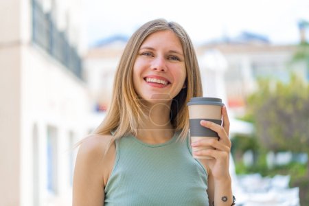 Photo for Young blonde woman at outdoors holding a take away coffee with happy expression - Royalty Free Image