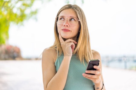 Téléchargez les photos : Jeune femme blonde à l'extérieur en utilisant le téléphone mobile et en pensant - en image libre de droit