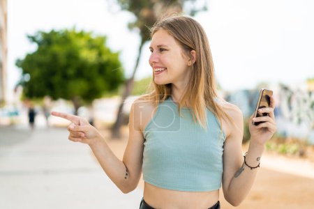 Foto de Mujer rubia joven utilizando el teléfono móvil al aire libre apuntando a un lado para presentar un producto - Imagen libre de derechos