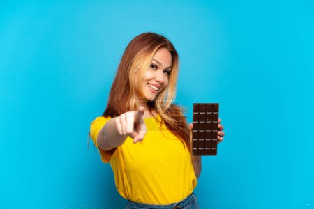 Foto de Adolescente chica sosteniendo chocolate sobre aislado fondo azul apuntando frente con expresión feliz - Imagen libre de derechos