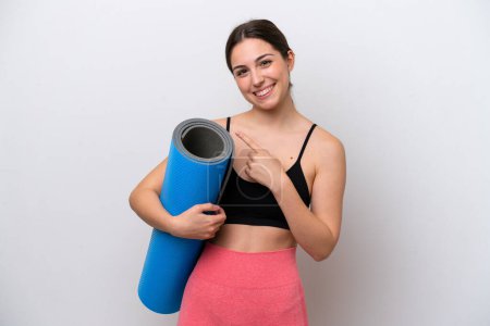 Téléchargez les photos : Jeune fille sportive allant à des cours de yoga tout en tenant un tapis isolé sur fond blanc pointant vers le côté pour présenter un produit - en image libre de droit