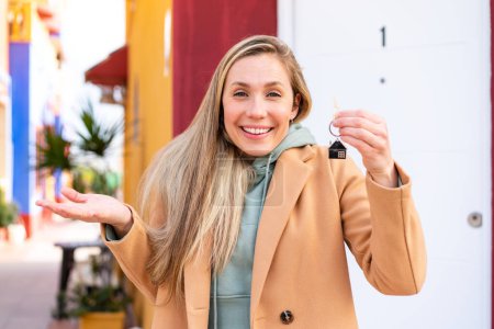 Téléchargez les photos : Jeune femme blonde tenant les clés de la maison à l'extérieur avec une expression faciale choquée - en image libre de droit