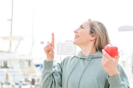 Foto de Mujer rubia joven con una manzana al aire libre con la intención de realizar la solución mientras levanta un dedo - Imagen libre de derechos