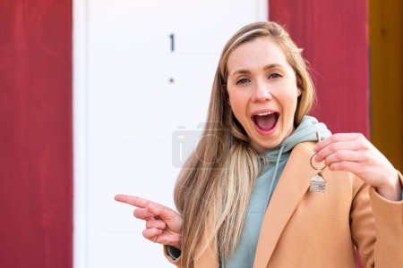 Foto de Joven mujer rubia sosteniendo las llaves de casa al aire libre sorprendida y señalando con el dedo hacia un lado - Imagen libre de derechos