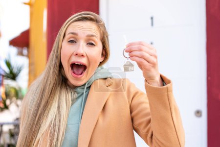 Téléchargez les photos : Jeune femme blonde tenant les clés de la maison à l'extérieur avec surprise et expression faciale choquée - en image libre de droit
