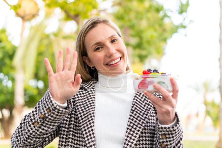 Photo for Young blonde woman holding a three dimensional puzzle cube at outdoors saluting with hand with happy expression - Royalty Free Image