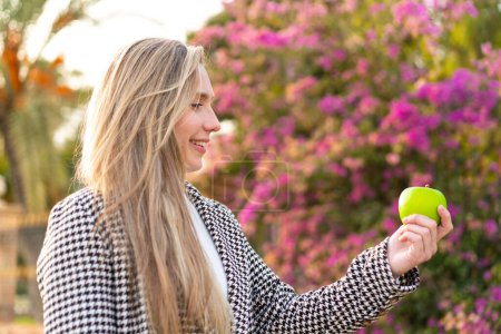 Foto de Mujer rubia joven con una manzana al aire libre con expresión feliz - Imagen libre de derechos