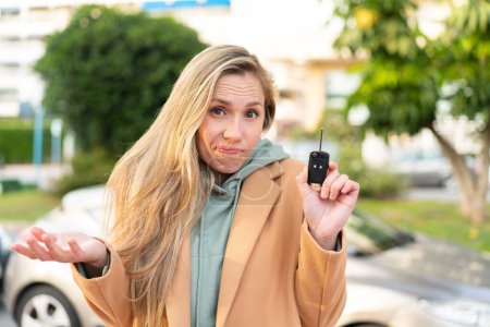 Foto de Joven mujer rubia sosteniendo las llaves del coche al aire libre haciendo gestos de dudas mientras levanta los hombros - Imagen libre de derechos