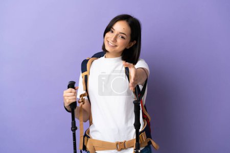 Foto de Mujer joven caucásica con mochila y bastones de trekking aislados sobre fondo azul estrechando las manos para cerrar una buena oferta - Imagen libre de derechos