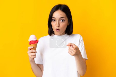 Foto de Mujer joven caucásica con un helado de corneta aislado sobre fondo amarillo sorprendido y apuntando hacia delante - Imagen libre de derechos