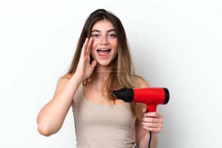 Téléchargez les photos : Jeune femme caucasienne tenant un sèche-cheveux isolé sur fond blanc criant avec la bouche grande ouverte - en image libre de droit