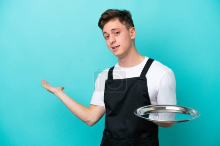 Photo for Young waitress with tray isolated on blue background extending hands to the side for inviting to come - Royalty Free Image