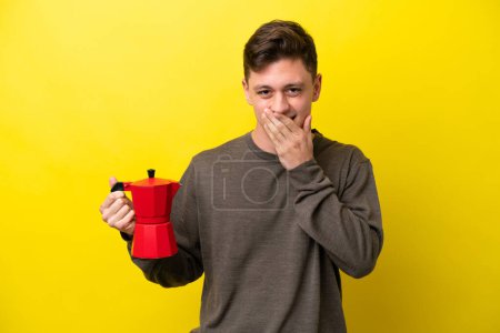 Foto de Joven brasileño sosteniendo cafetera aislada sobre fondo amarillo alegre y sonriente cubriendo boca con mano - Imagen libre de derechos