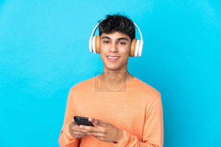 Foto de Joven sobre fondo azul aislado escuchando música con un móvil y mirando al frente - Imagen libre de derechos