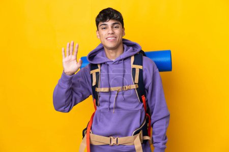 Téléchargez les photos : Jeune alpiniste homme avec un grand sac à dos sur fond jaune isolé saluant à la main avec expression heureuse - en image libre de droit