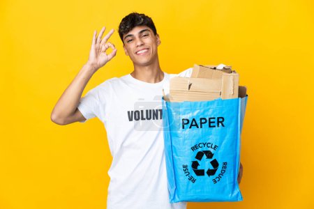 Téléchargez les photos : Homme tenant un sac de recyclage plein de papier à recycler sur fond jaune isolé montrant ok signe avec les doigts - en image libre de droit