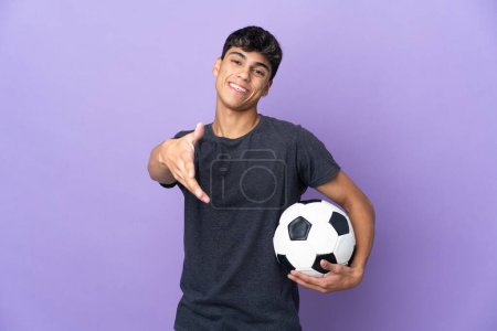 Foto de Joven jugador de fútbol hombre sobre fondo púrpura aislado estrechando las manos para cerrar una buena oferta - Imagen libre de derechos