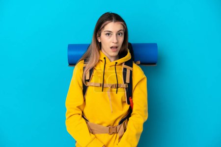 Foto de Young mountaineer woman with a big backpack isolated on blue background with surprise facial expression - Imagen libre de derechos