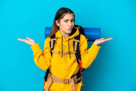 Photo for Young mountaineer woman with a big backpack isolated on blue background having doubts while raising hands - Royalty Free Image