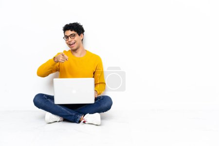 Téléchargez les photos : Homme vénézuélien assis sur le sol avec ordinateur portable surpris et pointant devant - en image libre de droit