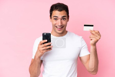 Photo for Young caucasian man isolated on pink background buying with the mobile and holding a credit card with surprised expression - Royalty Free Image