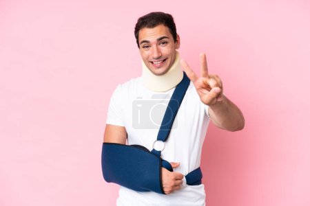 Téléchargez les photos : Jeune homme caucasien avec le bras cassé et portant une fronde isolée sur fond rose souriant et montrant signe de victoire - en image libre de droit