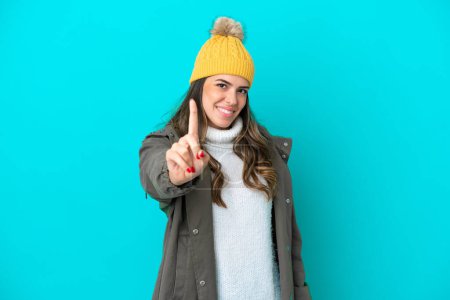 Téléchargez les photos : Jeune femme italienne vêtue d'une veste d'hiver et chapeau isolé sur fond bleu montrant et levant un doigt - en image libre de droit