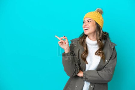 Téléchargez les photos : Jeune femme italienne vêtue d'une veste d'hiver et chapeau isolé sur fond bleu pointant du doigt vers le côté et présentant un produit - en image libre de droit