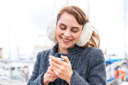Photo for Brunette woman wearing winter muffs at outdoors sending a message with the mobile - Royalty Free Image